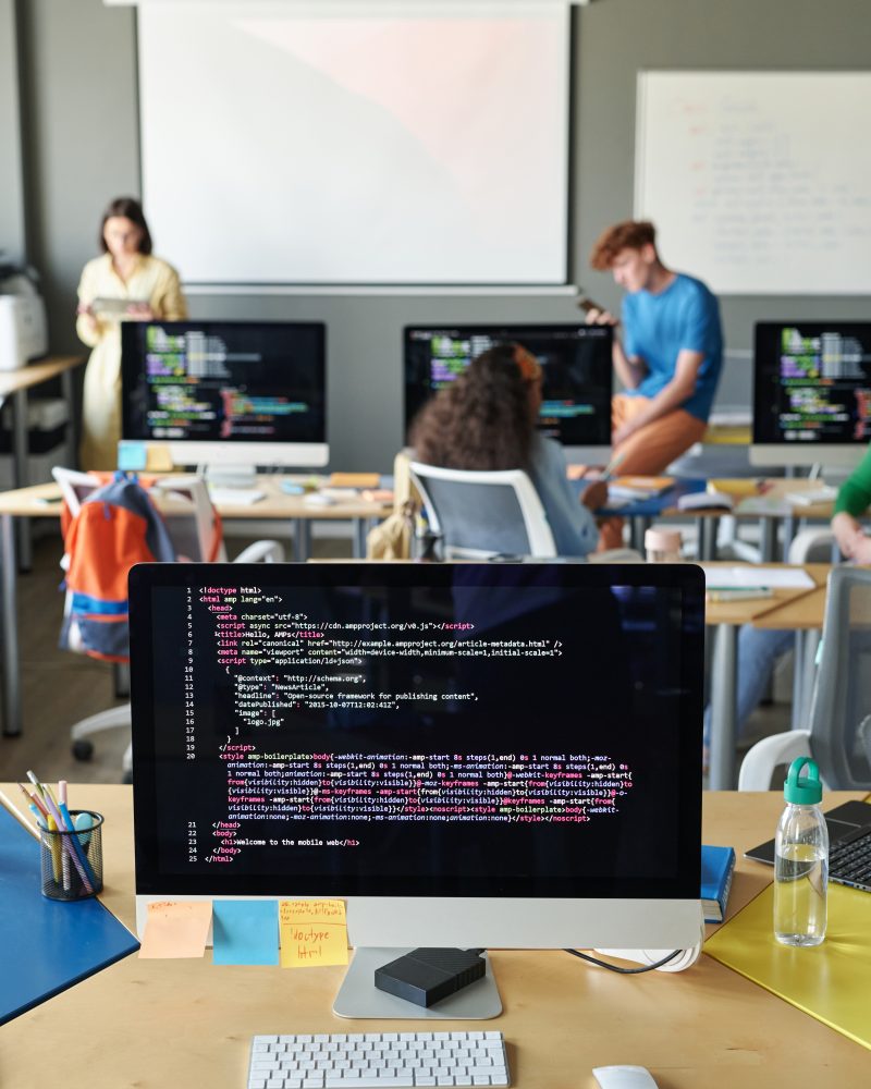 Vertical image of computer with codes on monitor standing on desk in the classroom with students and teacher in background